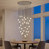 A silver-toned pendant chandelier with glowing tube ends hangs above a round wooden dining table beside tall windows.