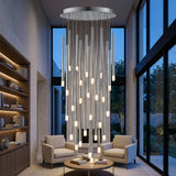 A silver-toned pendant chandelier hangs above a round coffee table between cream armchairs, beside illuminated shelves and tall windows.