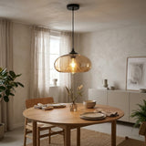A modern pendant light with a translucent amber-tinted glass dome and a black ceiling canopy, hanging above a round wooden dining table in a softly lit, neutral-toned dining room.