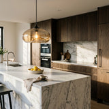 A modern kitchen interior with a large white marble island, dark wood cabinets, a glass pendant light, and countertop decor including lemons and a cup on a saucer.