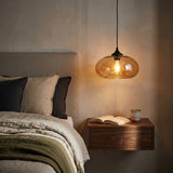 A modern pendant light with a rounded amber-tinted glass shade and a black cord, hanging above a floating wooden nightstand in a bedroom setting.