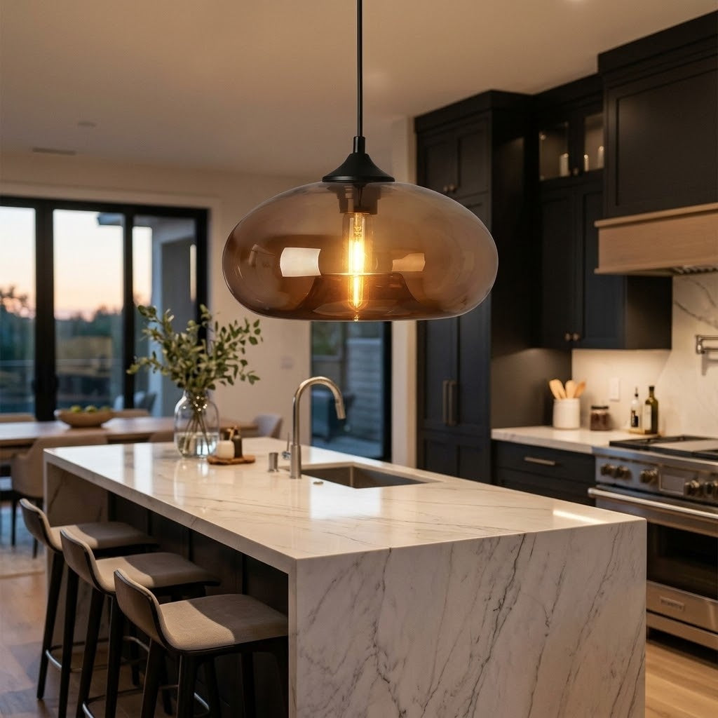 A modern kitchen island with a large oval amber-tinted glass pendant light hanging above it. The kitchen features dark charcoal cabinets, a white marble island, and a built-in gas stove in the background.