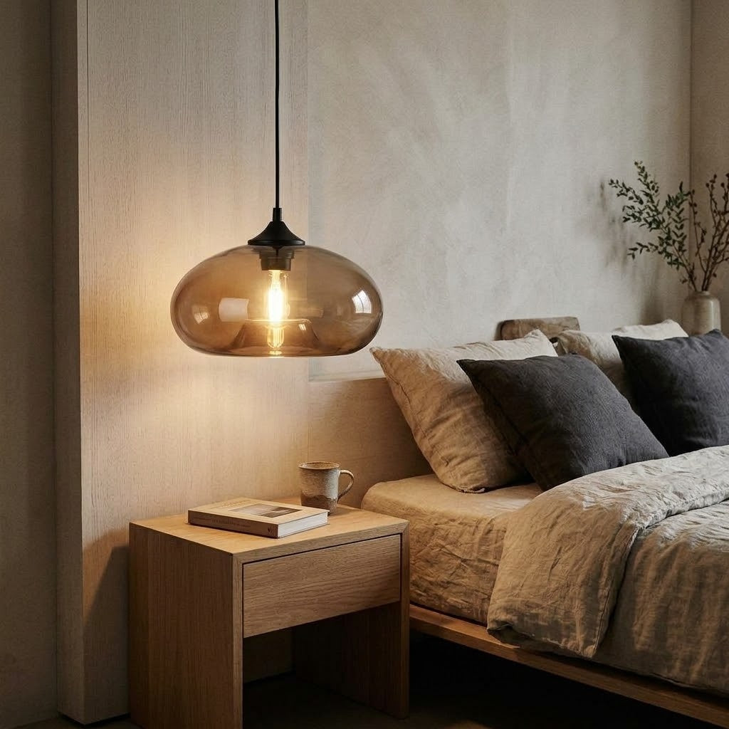 A modern pendant light with a translucent amber glass dome and a visible warm bulb, hanging above a wooden bedside table in a softly lit bedroom with a bed and neutral-toned bedding.