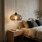 A modern pendant light with a translucent amber glass dome and a visible warm bulb, hanging above a wooden bedside table in a softly lit bedroom with a bed and neutral-toned bedding.