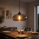 A warm amber glass pendant light hangs over a wooden dining table set with plates, wine glasses, and a small vase in a softly lit dining room.