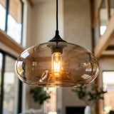 A modern pendant light with a large, rounded translucent amber-tinted glass shade and a visible glowing filament bulb, hanging in a softly blurred interior setting.