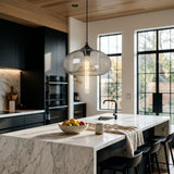 A modern kitchen interior with a large glass pendant light hanging over a marble island. The scene includes black cabinetry, large black-framed windows, a bowl of fruit on the island, a mug, and black bar stools.
