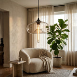 A modern pendant light with a clear glass dome shade and visible Edison-style bulb, hanging in a softly lit living room with a cream boucle armchair, sheer curtains, and a large green plant.