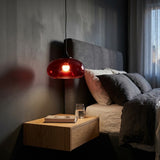 Red oval glass pendant light with a glowing bulb hangs above a floating wooden nightstand beside a bed with a grey upholstered headboard.