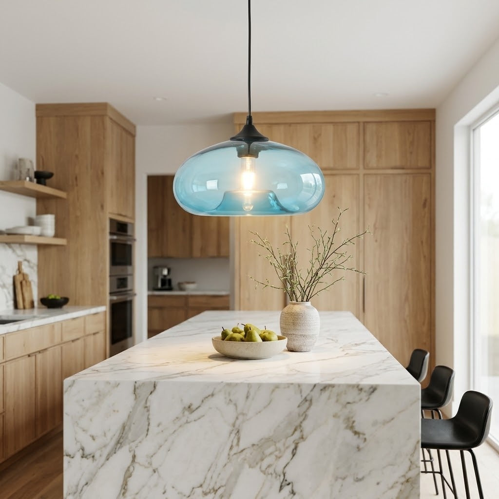 A modern kitchen scene with a blue glass pendant light hanging over a marble island. The kitchen features light wood cabinets, a white marble island with gray veining, a small vase with dried branches, and a bowl of green pears.