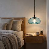 A modern pendant light with a translucent blue-green glass dome shade hanging over a wooden nightstand in a bedroom setting.