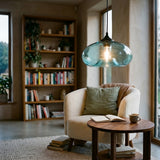 A modern pendant light with a translucent blue-green glass dome shade hangs over a cozy reading nook with a cream bouclé armchair, wooden bookshelf, and round coffee table.