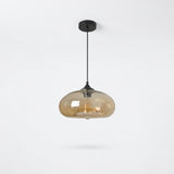 A modern pendant light with a black ceiling canopy and cord, featuring a translucent amber-tinted glass shade that reveals a warm glowing filament bulb inside, set against a clean white background.