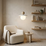 A modern pendant light with a white conical shade and a warm beige spherical accent hangs above a cream bouclé armchair in a softly styled room with floating wooden shelves, a small side table, and a mug.