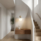 A modern pendant light with a spherical wooden top and glowing white globe shade, hanging in a minimalist staircase hallway with light wood floors and white walls.