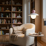 A cozy living room scene featuring a cream bouclé armchair with a beige throw blanket, set in front of a tall wooden bookshelf. A small marble-topped side table with a mug and open book sits nearby under a modern pendant light with a pink top and white globe shade.