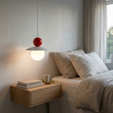 A modern pendant light with a red spherical top and a glowing white globe shade, hanging above a minimalist wooden bedside table in a softly lit bedroom.
