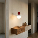 A modern pendant light with a white conical shade, a glowing spherical bulb, and a glossy red top sphere, hanging in a minimalist interior above a wooden console table.