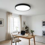 A modern circular ceiling light with a matte black frame and bright white diffuser, mounted on a white ceiling in a bright, minimalist home office.