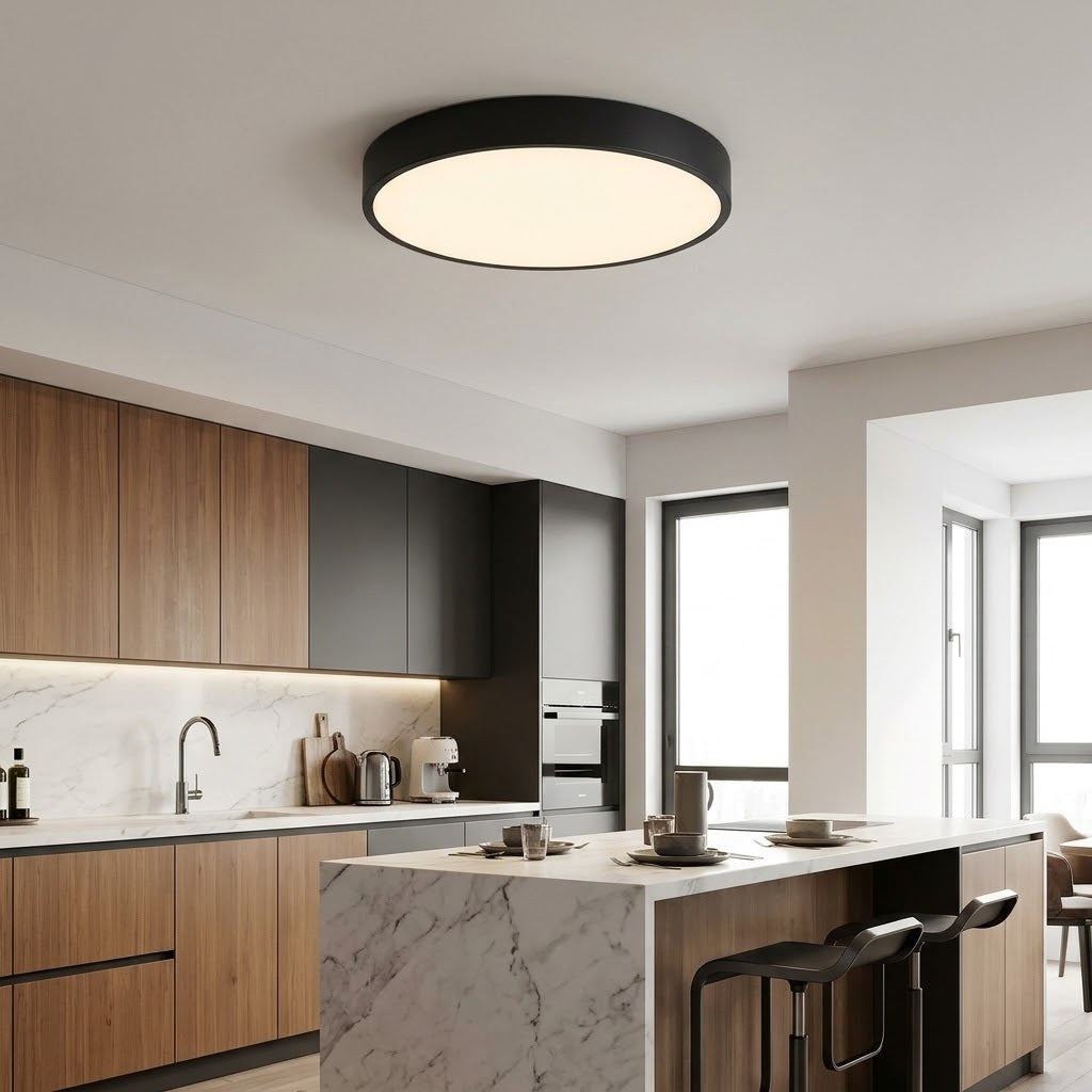 A modern circular ceiling light with a matte black frame and warm white glowing diffuser, mounted on the ceiling of a contemporary kitchen with wood and dark gray cabinets.