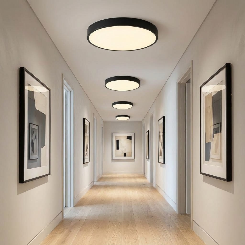 A hallway with light wood flooring and beige walls, illuminated by three modern circular ceiling lights with black frames and warm white glowing diffusers. Framed abstract wall art hangs on both sides.