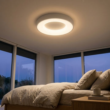 A modern circular ceiling light with a warm white glow installed on the ceiling of a bedroom, with a bed and large windows in the background.