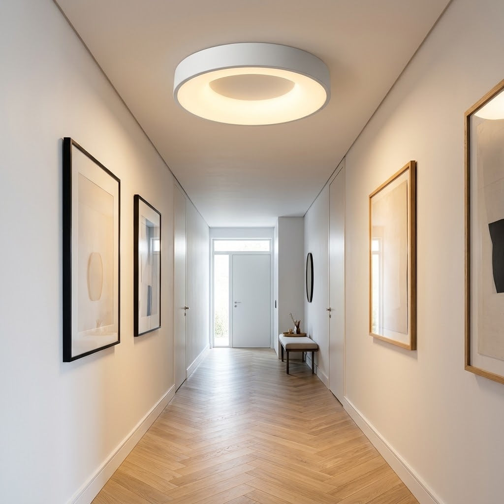 A modern circular flush-mount ceiling light with a white outer rim and a warm white glowing inner ring, installed in a bright hallway with light wood herringbone flooring and framed wall art.