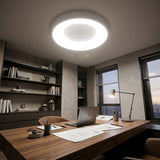 A modern circular LED ceiling light glowing brightly in a contemporary home office with a wooden desk, laptop, and shelves in the background.