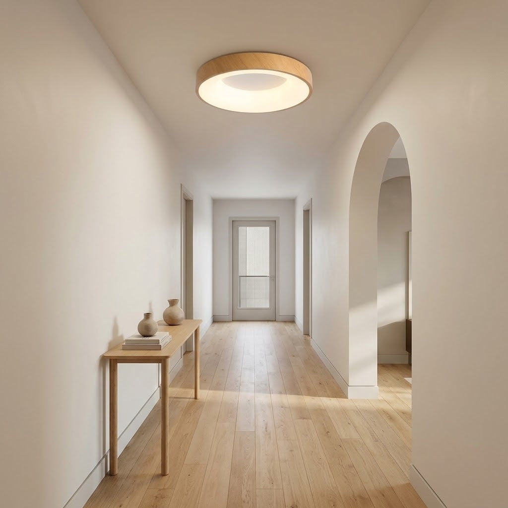 A modern circular flush-mount ceiling light with a light wood rim and glowing white diffuser, installed in a minimalist hallway with light wood floors and white walls.