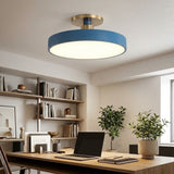 A modern semi-flush mount ceiling light with a blue circular shade and a brushed gold metal neck, installed in a bright, contemporary home office with a wooden desk, laptop, and plants.