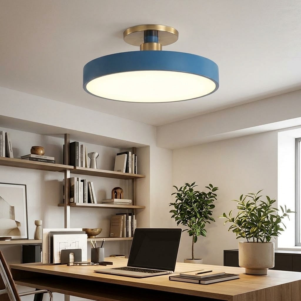 A modern semi-flush mount ceiling light with a blue circular shade and a brushed gold metal neck, installed in a bright, contemporary home office with a wooden desk, laptop, and plants.