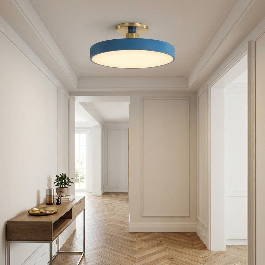 A modern semi-flush mount ceiling light with a blue cylindrical shade and a brushed gold brass ceiling mount, installed in a bright, elegant hallway with cream walls and herringbone wood floors.