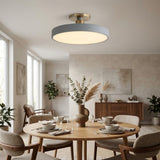 A modern semi-flush mount ceiling light with a matte blue-gray circular shade and a brushed brass ceiling mount, shown lit in a beautifully styled dining room with a wooden dining table, chairs, and neutral decor.