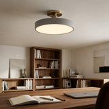 A modern semi-flush mount ceiling light with a matte gray circular shade and a brushed brass ceiling mount, shown illuminated in a warm, contemporary home office setting with a wooden desk, laptop, and bookshelves.