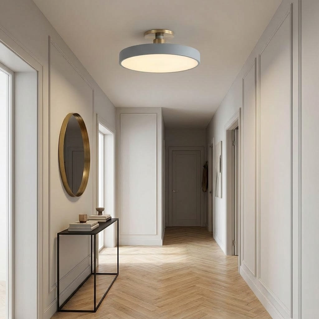 A modern flush-mount ceiling light with a circular light gray shade and a brushed gold metal frame, installed in a bright, elegant hallway with light wood herringbone floors and white paneled walls.