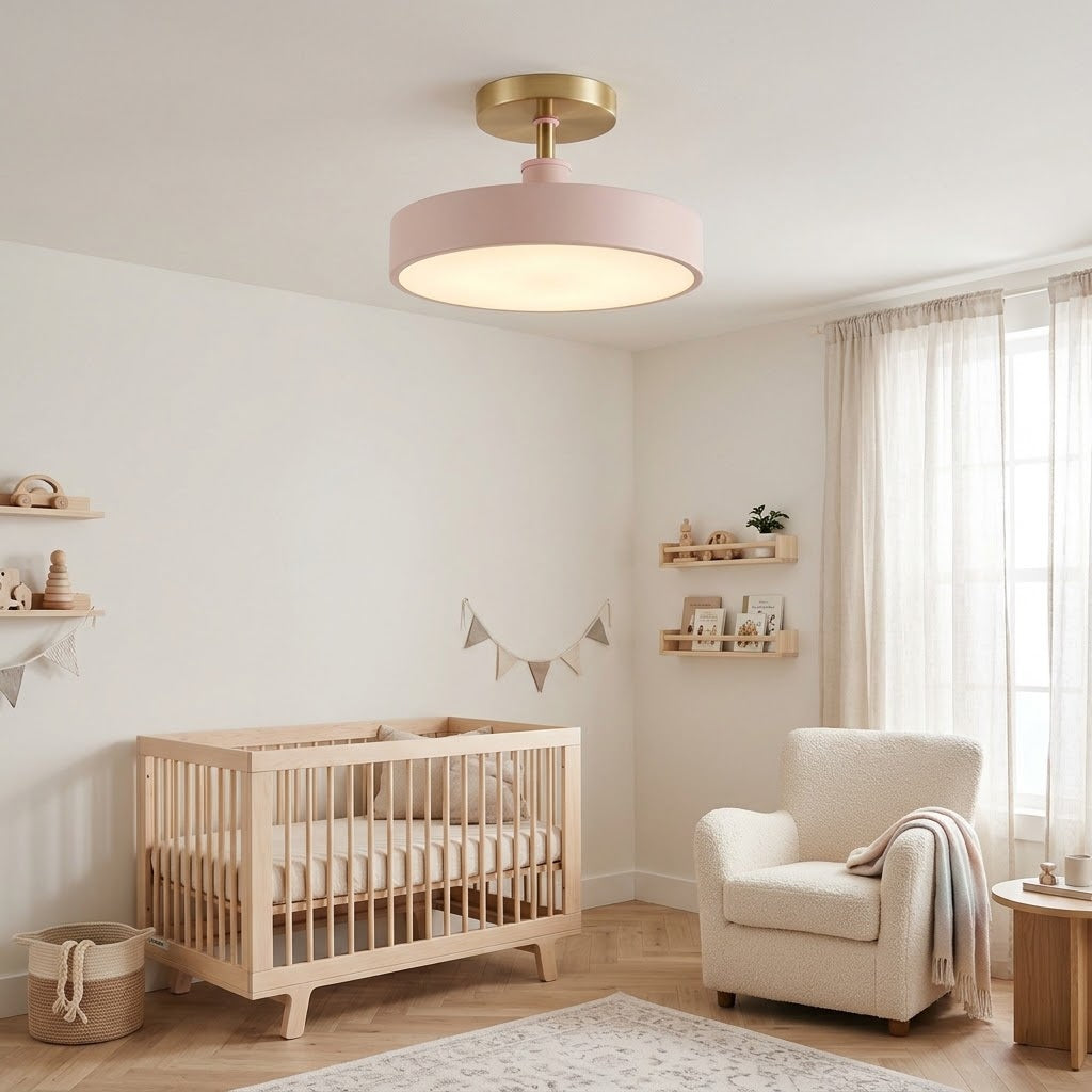 A modern ceiling light fixture with a round blush pink shade and a gold-toned ceiling mount, shown installed in a softly styled nursery with a crib, armchair, and curtains.
