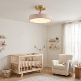 A modern ceiling light fixture with a round blush pink shade and a gold-toned ceiling mount, shown installed in a softly styled nursery with a crib, armchair, and curtains.