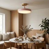 A modern semi-flush mount ceiling light with a matte pink circular shade and a brushed brass gold ceiling mount, hanging over a styled dining table in a warm, minimalist room.