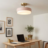 A modern semi-flush mount ceiling light with a matte blush pink cylindrical shade and a brushed brass ceiling mount, shown illuminated in a styled home office setting with a wooden desk, laptop, and potted plant.