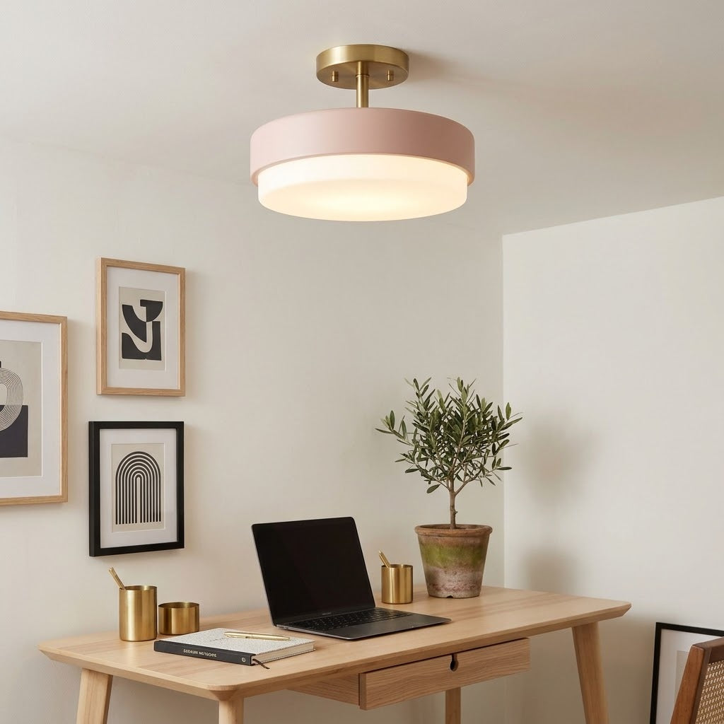 A modern semi-flush mount ceiling light with a matte blush pink cylindrical shade and a brushed brass ceiling mount, shown illuminated in a styled home office setting with a wooden desk, laptop, and potted plant.