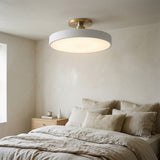 A modern semi-flush mount ceiling light with a matte white circular shade and a brushed gold brass ceiling mount, shown installed in a minimalist beige bedroom above a bed with neutral linens.