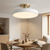 A modern semi-flush mount ceiling light with a white circular shade and a brushed gold metal ceiling mount, shown illuminated in a warm, contemporary dining room setting.