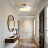 A modern flush-mount ceiling light with a white cylindrical shade and a brushed gold circular backplate, mounted in a bright, elegant white hallway with wood floors and a black front door.