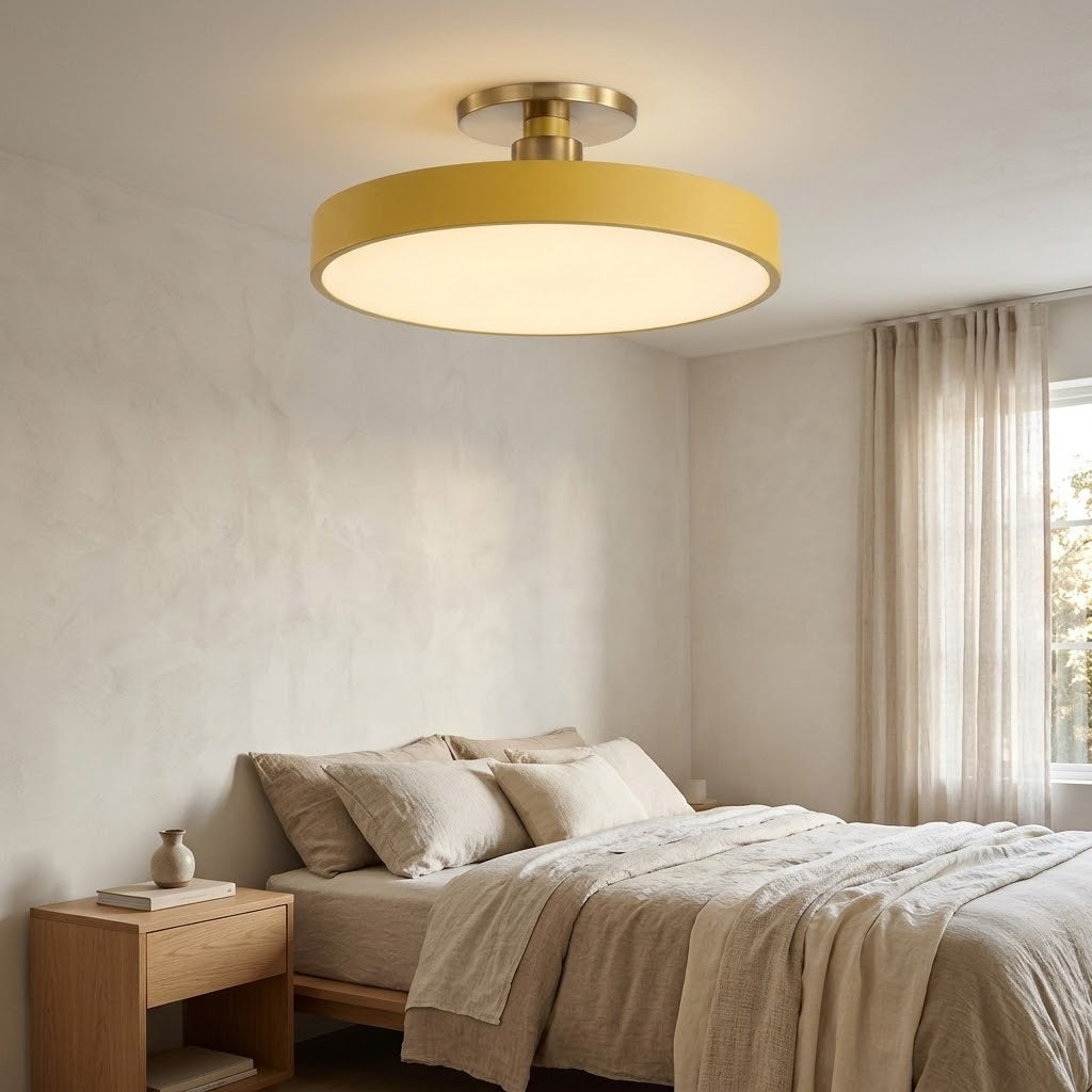 A modern semi-flush mount ceiling light with a wide, circular drum shade in a warm mustard yellow and a brushed brass metal top canopy, installed in a minimalist beige bedroom.