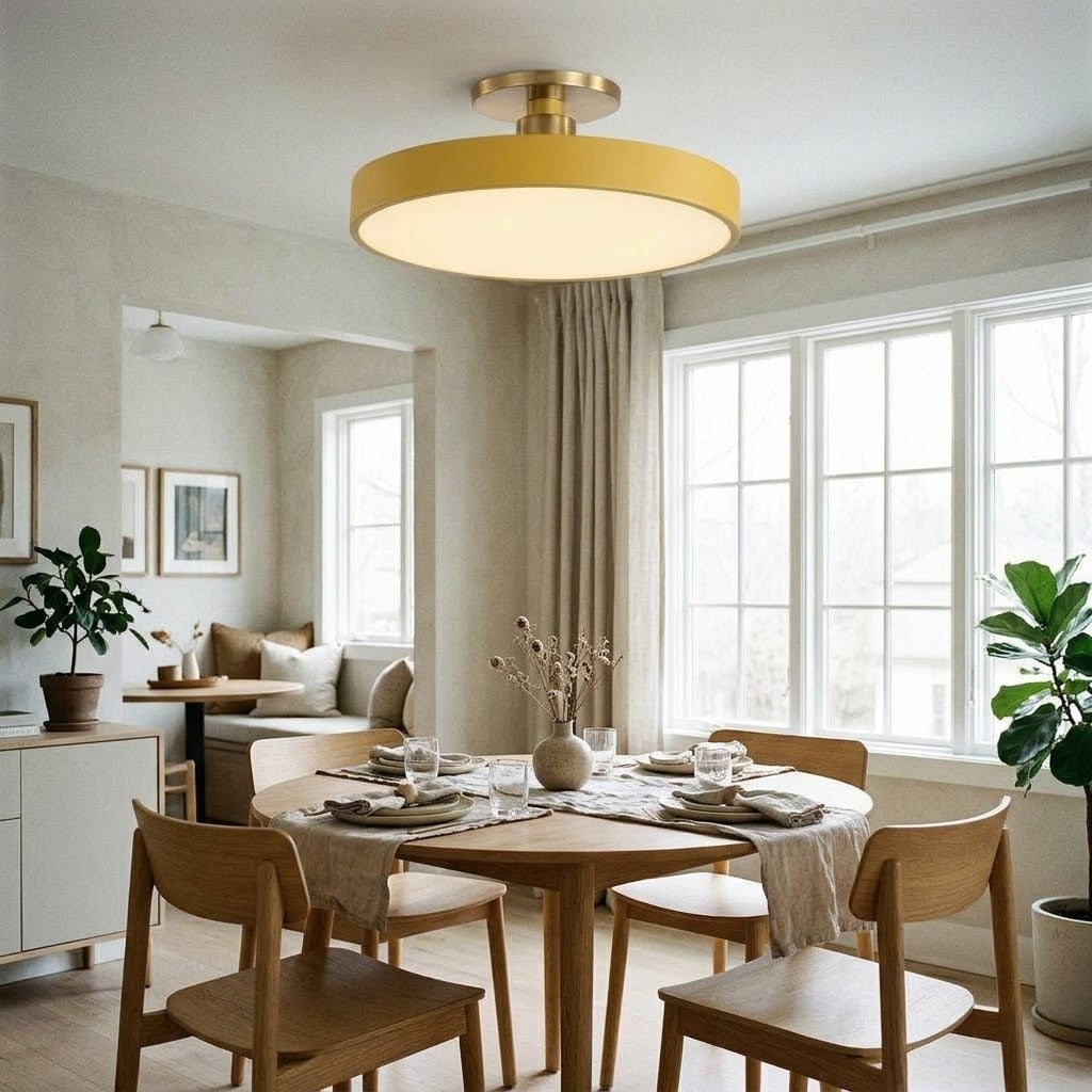 A modern semi-flush mount ceiling light with a circular shade and gold-toned metal hardware, installed in a bright, contemporary dining room with wooden chairs and a round table.