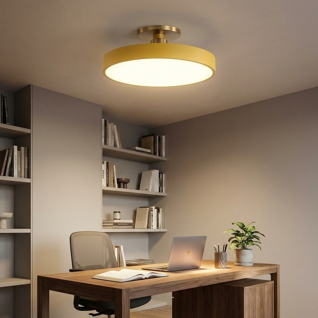 A modern semi-flush mount ceiling light with a circular warm yellow shade and gold-toned hardware, installed in a contemporary home office setting above a wooden desk.