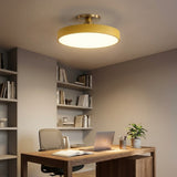 A modern semi-flush mount ceiling light with a circular warm yellow shade and gold-toned hardware, installed in a contemporary home office setting above a wooden desk.