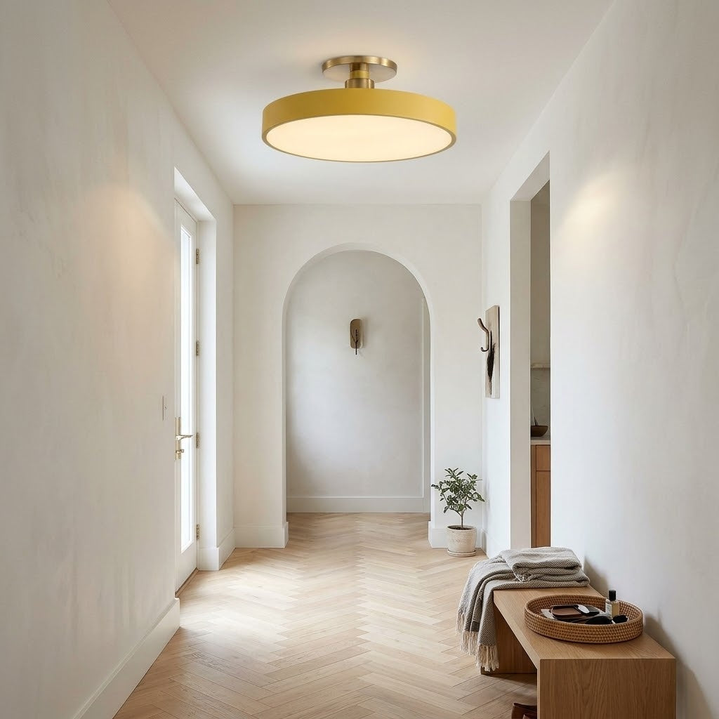 A modern circular flush-mount ceiling light with a warm gold-toned frame and glowing white glass, installed in a bright, minimalist hallway with light wood floors and white walls.