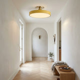 A modern circular flush-mount ceiling light with a warm gold-toned frame and glowing white glass, installed in a bright, minimalist hallway with light wood floors and white walls.