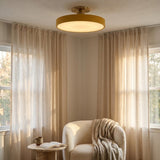 A round flush-mount ceiling light with a warm brass/gold-toned frame and glowing warm white light, installed in a cozy room corner with beige curtains, a cream armchair, and a small side table.